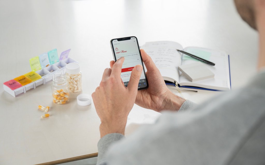 Hands holding a mobile phone with screen showing a Zur Rose page. Pills, pill organizer and a notebook are in the background