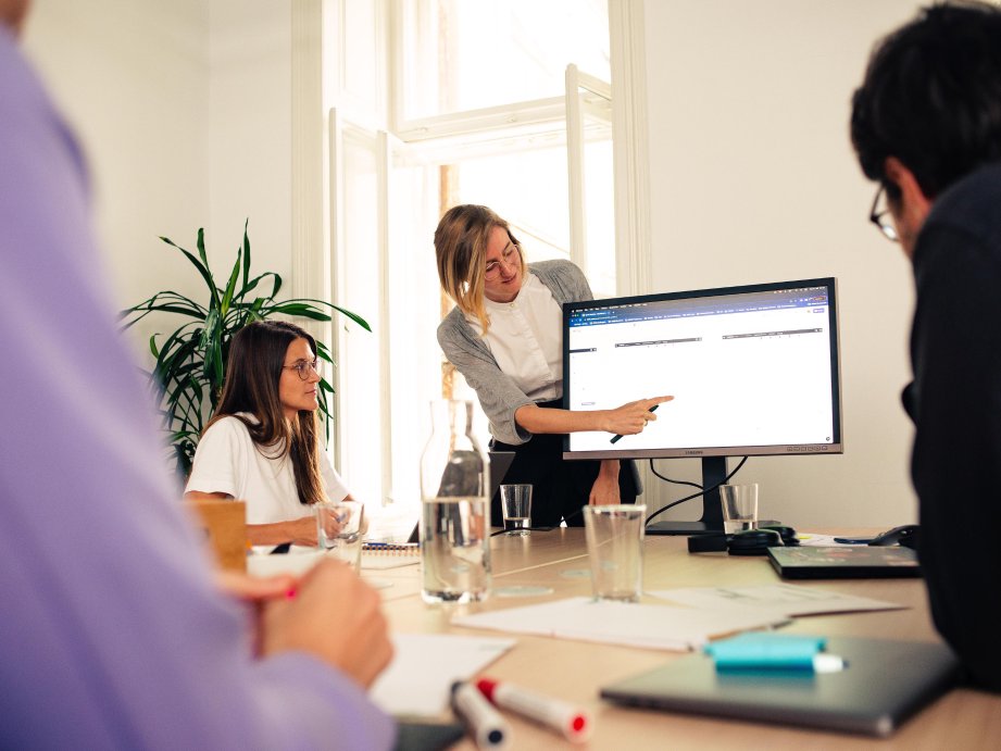 Three people sitting at a table during client requirement workshop while a woman presents, and points at a screen